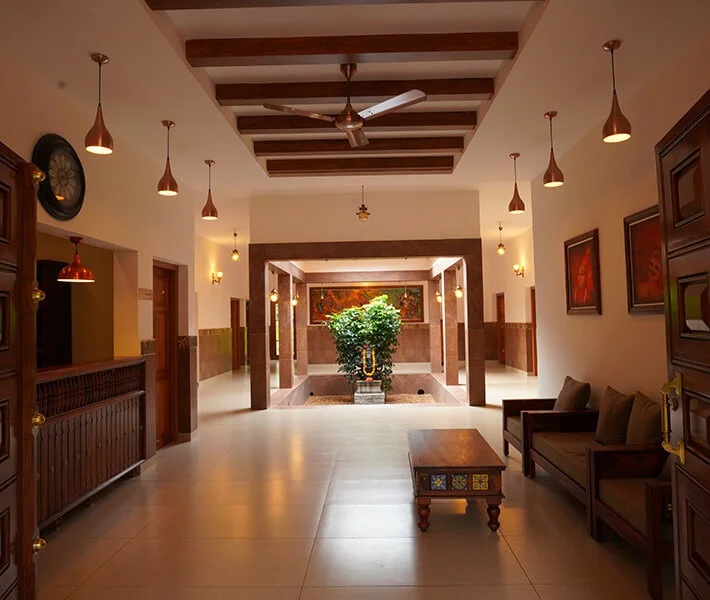 Warm, elegant hotel lobby with wood beams, pendant lights, ceiling fan, tiled floor, carved coffee table, sofas, and a leafy courtyard plant.