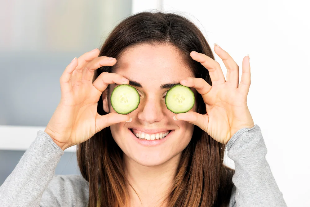 Smiling woman in a gray sweater holds cucumber slices over her eyes in a playful, refreshing skincare treatment in a bright room.
