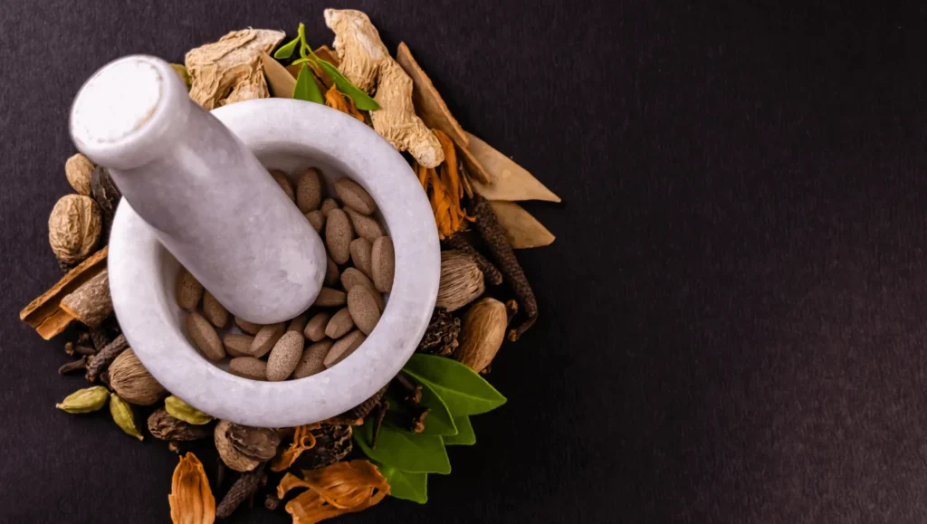 Overhead view of a white mortar and pestle filled with almonds, surrounded by assorted spices and herbs on a dark rustic background.