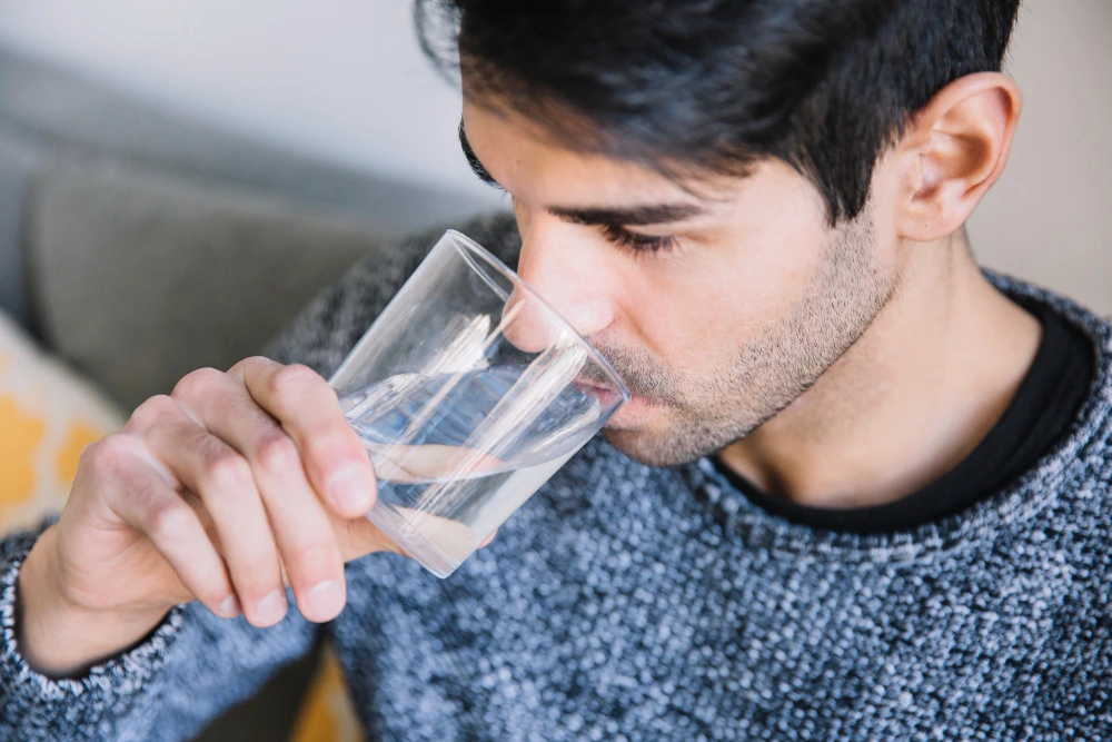 Close-up of a young man in a gray sweater drinking water from a clear glass, creating a calm, everyday moment indoors.