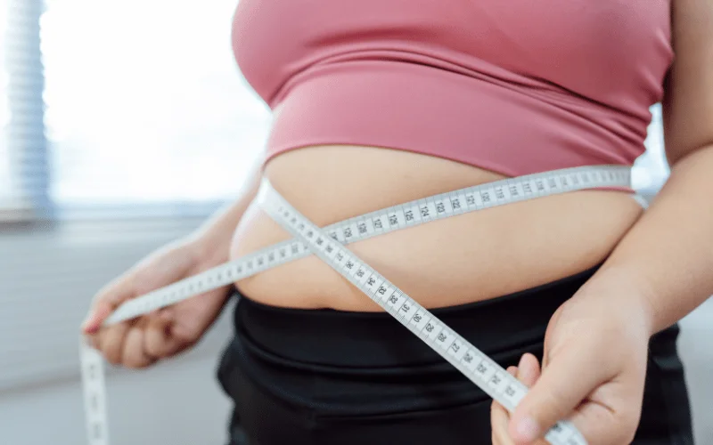 Close-up of a person in a pink top and black pants measuring their waist with a tape measure, suggesting health and weight-loss goals.