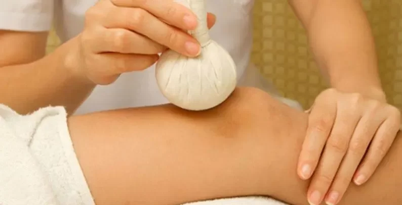 Close-up of a therapist applying a warm herbal compress to a client’s leg during a soothing spa treatment, with white towels visible.