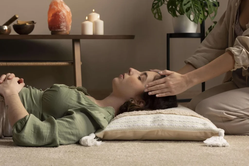 A serene woman lies on a cushion with eyes closed while another gently cradles her head, creating a calming, restorative wellness scene.