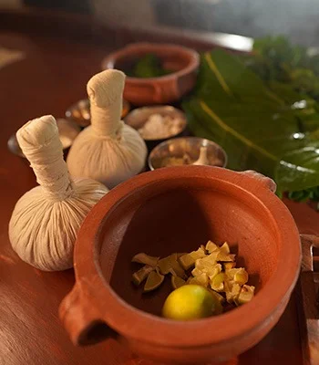 Warm still life of a clay mortar with lime and dried herbs, surrounded by tied herbal sachets, bowls, and fresh green leaves on wood.