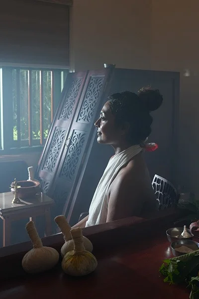 Serene woman in profile seated indoors beside a carved decorative screen, sunlit window, herbal bowls, and small gourds on a table.