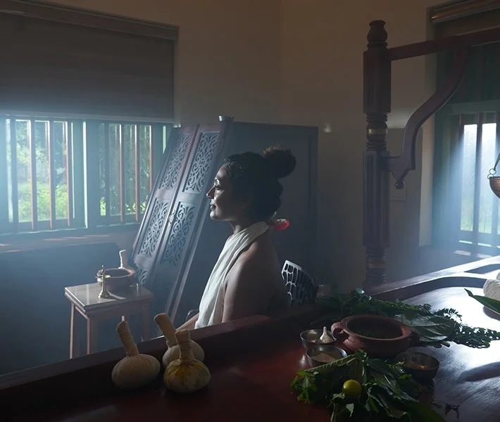 A serene woman sits in a sunlit wooden room beside herbal compress balls, bowls, greenery, and traditional furnishings, creating a tranquil mood.
