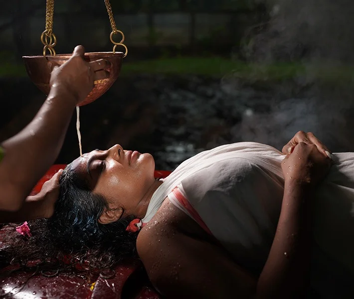 A serene woman in a white sari reclines by a stream as water pours from a suspended copper vessel during a bathing ritual.