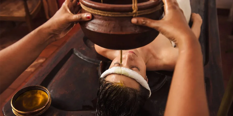 Ayurvedic oil treatment: a therapist pours warm oil from a wooden vessel onto a client's forehead as they relax, conveying calm and wellness.