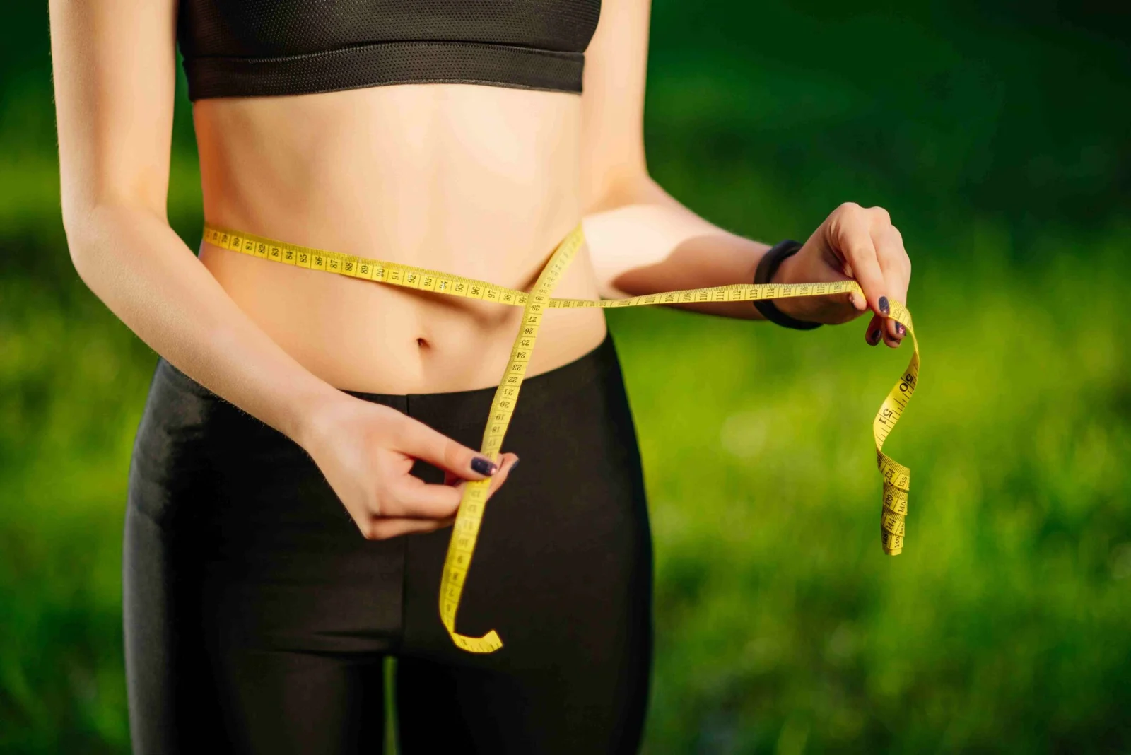 Slim person in black workout clothes measures their waist with a yellow tape measure outdoors, conveying fitness, health, and body transformation.