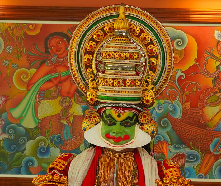 Kathakali performer in vivid green makeup and ornate headdress poses before a colorful mural, conveying ceremonial drama and cultural richness.