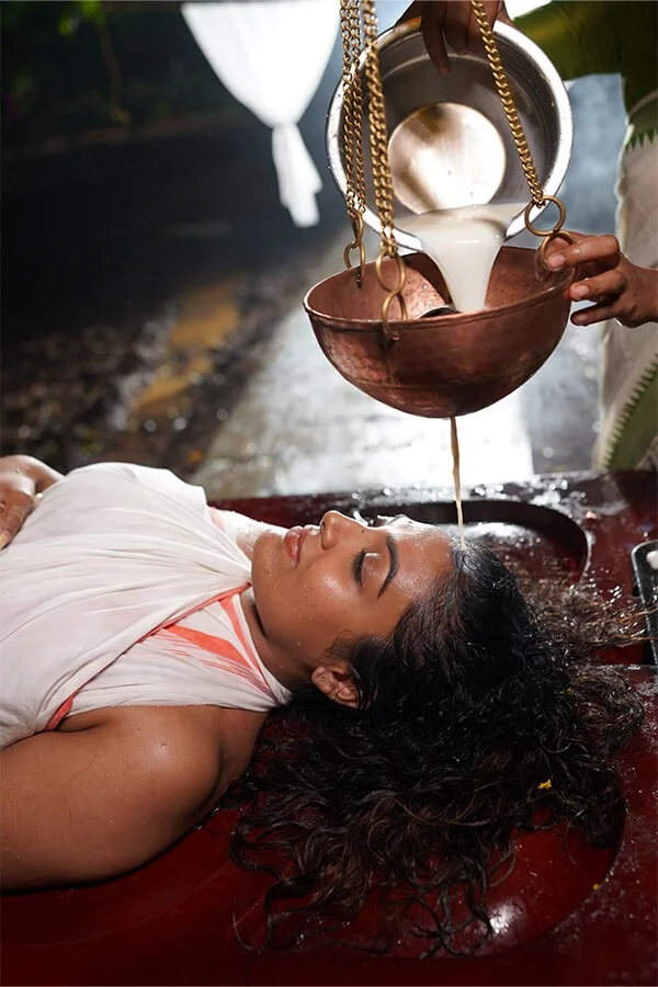 A serene woman lies with eyes closed as a suspended brass vessel pours white liquid over her forehead during a peaceful wellness ritual.