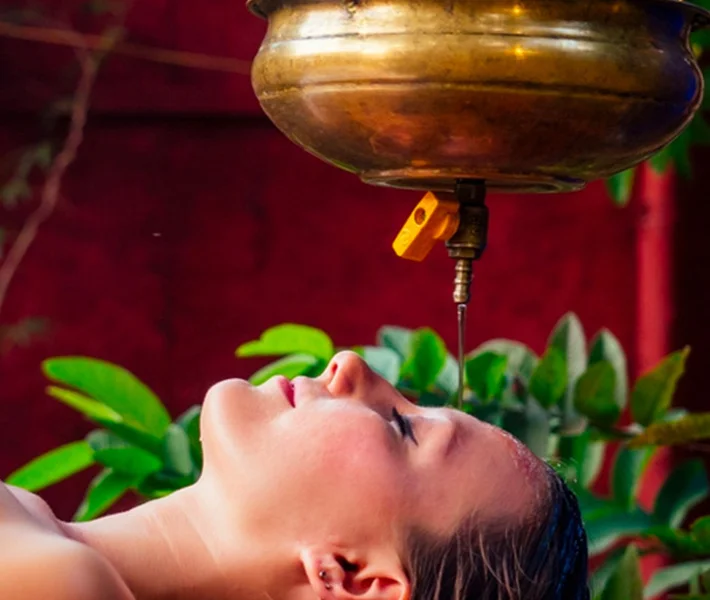 Woman lies with eyes closed beneath a brass Ayurvedic shirodhara vessel as warm oil drips onto her forehead in a serene spa setting.