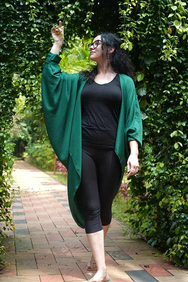Woman in glasses, black outfit, and flowing green cardigan walks barefoot on a leafy garden path, pointing upward with a relaxed expression.