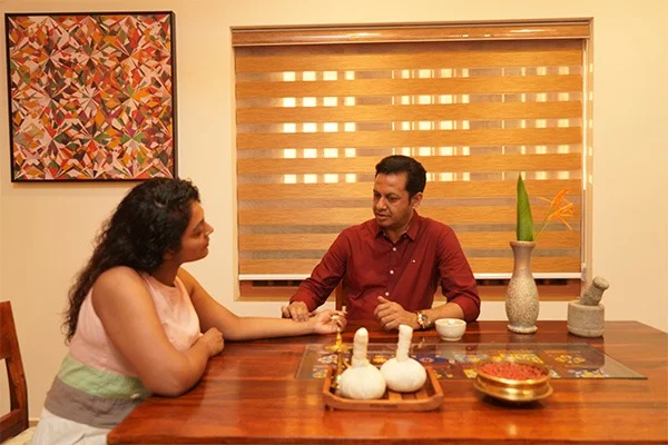 A woman and man converse warmly at a dining table, surrounded by candles, a bowl, plants, artwork, and wooden window blinds.