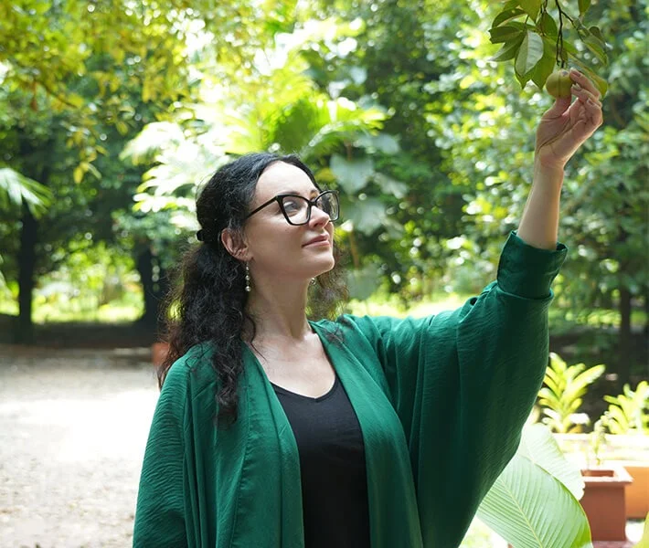 Woman in glasses reaches toward a leafy branch in a sunlit garden, wearing green; her calm, thoughtful expression creates a serene mood.