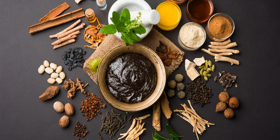 Overhead view of a bowl of dark spice paste surrounded by cinnamon, herbs, nuts, seeds, dried chilies, and colorful spices on a rustic table.