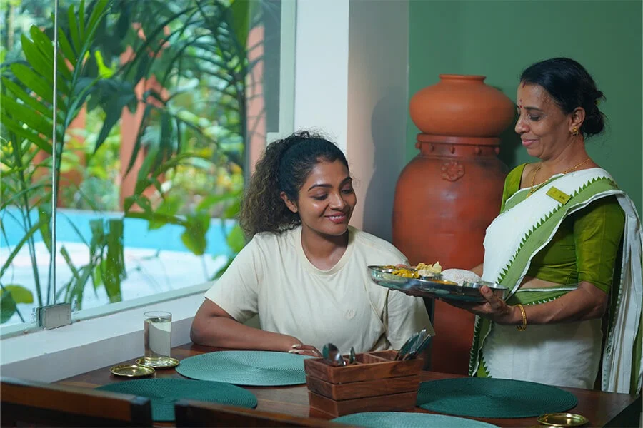 A smiling woman serves food to a seated young woman at a dining table, in a bright home with plants and a terracotta pot.