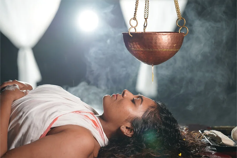 A woman lies peacefully beneath a suspended copper bowl as smoke swirls around her, creating a mystical, ritual-like atmosphere.