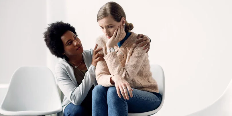 Two women sit closely in white chairs; one comforts the other, who looks sad and withdrawn, conveying empathy, support, and emotional care.