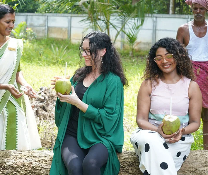 Three smiling women hold fresh coconuts outdoors, joined by a man in traditional clothing; lush greenery creates a warm, joyful setting.