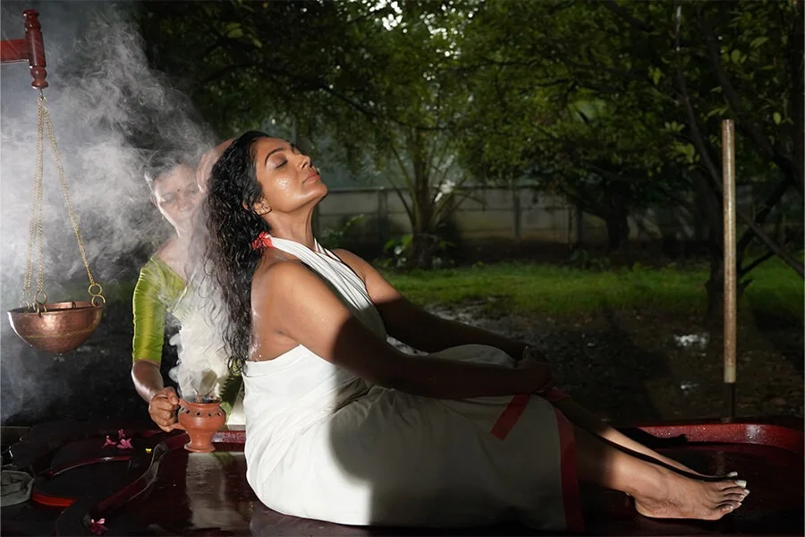 A woman in a white sari sits peacefully outdoors as incense smoke rises from a brass bowl, with a person behind her and greenery beyond.