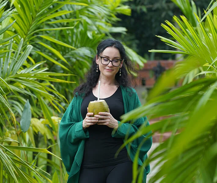Woman with glasses holds a fresh coconut and straw among lush tropical palm leaves, creating a serene, refreshing outdoor scene.
