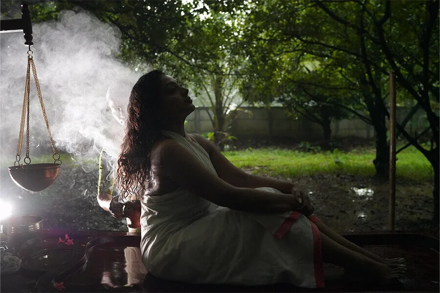 A woman in a white dress sits beside a steaming outdoor bath beneath trees, eyes closed in peaceful meditation as warm, misty light surrounds her.