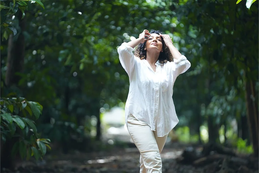 A smiling woman in a white blouse and beige pants walks through a lush forest, lifting her hands to her curly hair in bright, peaceful light.