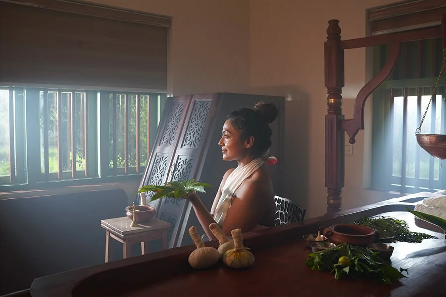Woman with a bun sits at a wooden table holding fresh herbs in a sunlit tropical home, surrounded by vegetables and woven decor.