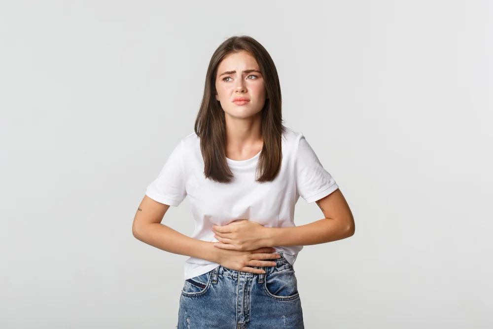 Young woman in a white T-shirt and jeans clutches her stomach, looking worried and uncomfortable against a plain gray background.