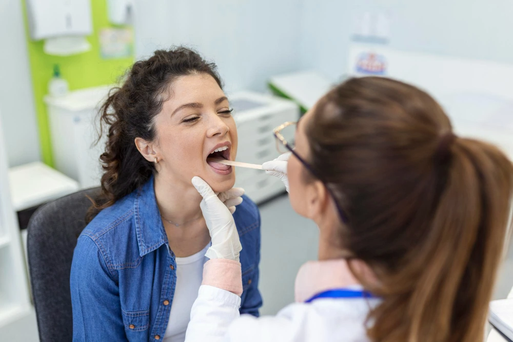 Dentist in gloves examines a smiling woman’s open mouth with a dental mirror in a bright clinic, creating a calm, professional scene.