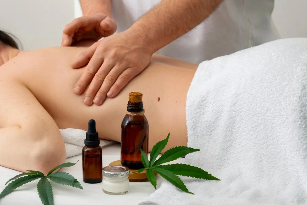 Massage therapist gently rubs a client’s back beside hemp leaves and amber oil bottles, creating a calm, soothing spa atmosphere.