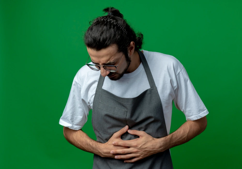 A worried man in glasses and a gray apron clutches his stomach, conveying abdominal pain or discomfort against a green background.