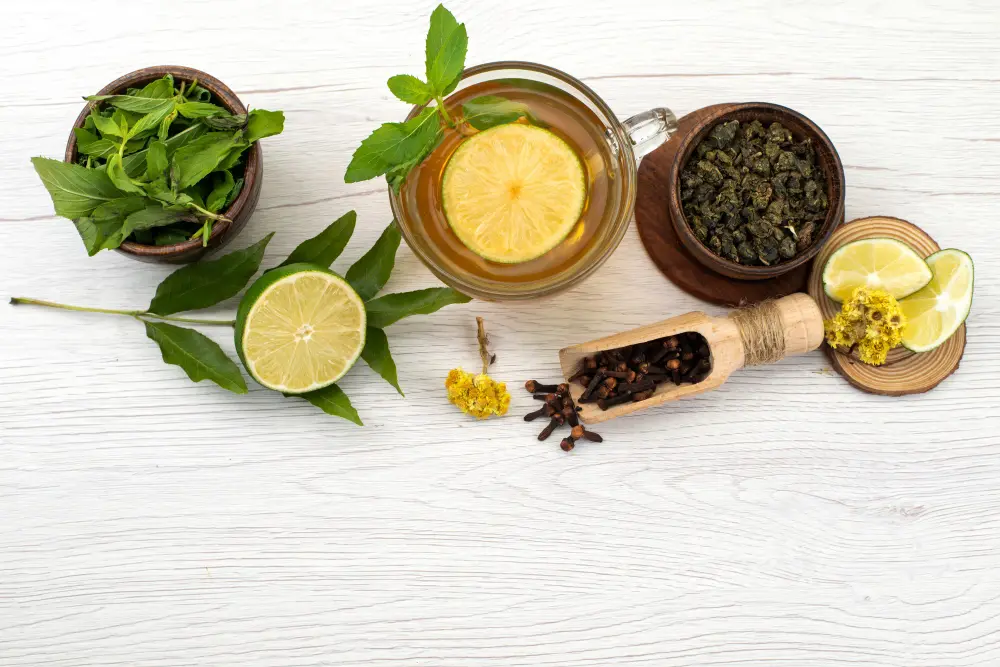 Overhead view of mint leaves, lime slices, herbal tea, dried tea leaves, spices, and lemon on a white wood table, creating a fresh, calming scene.